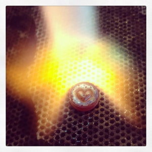 May include: A heart-shaped pendant is being heated with a torch on a metal grid. The pendant is surrounded by a bright orange glow.