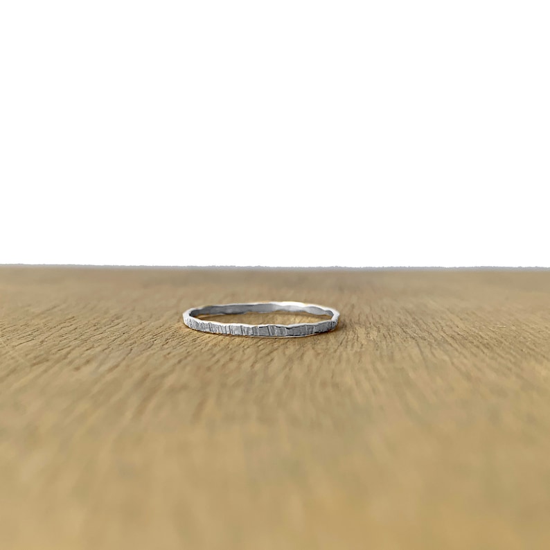 May include: A thin silver ring with a textured surface. The ring is lying on a wooden surface.