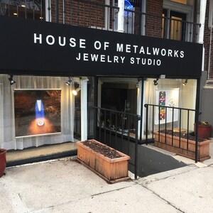 May include: A storefront with a black awning that reads "House of Metalworks Jewelry Studio". The storefront has two large windows, one displaying a piece of jewelry and the other displaying a collection of colorful stones. There are two wooden planters with plants in front of the store.