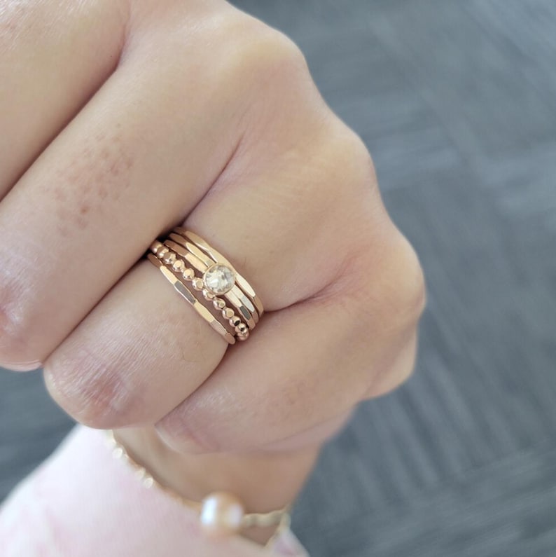 May include: A set of five gold-toned stacking rings with a small clear gemstone in the center. The rings are worn on a finger.