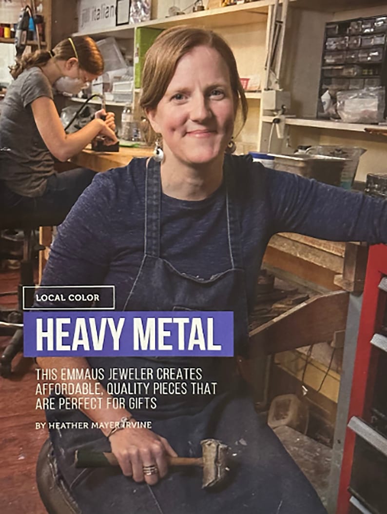 May include: A woman wearing a blue denim overall dress and a blue shirt is working in a jewelry studio. She is holding a hammer in her hand. The text "LOCAL COLOR HEAVY METAL" is displayed above her. The text "THIS EMMAUS JEWELER CREATES AFFORDABLE, QUALITY PIECES THAT ARE PERFECT FOR GIFTS BY HEATHER MAYER IRVINE" is displayed below her.
