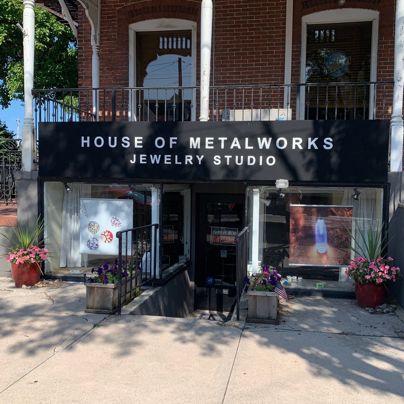 May include: A storefront with a black awning that reads "House of Metalworks Jewelry Studio". The storefront has two large windows with displays of jewelry inside. There are plants in pots outside the store.