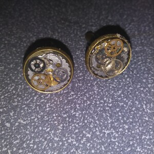 May include: A pair of gold cufflinks with a clear resin top. The resin contains a variety of small gears and cogs.