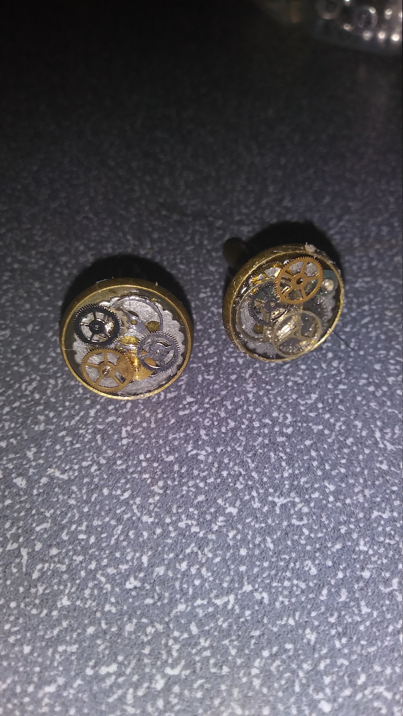 May include: A pair of gold cufflinks with a clear resin top. The resin contains a variety of small gears and cogs.