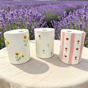May include: Three white ceramic vases with floral designs. One vase features sunflowers, another has small flowers, and the third has pink stripes with red hearts and flowers. The vases are cylindrical and sit on a beige surface.
