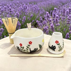 Könnte beinhalten: Ein weißes Keramik-Matcha-Schalen-, Schneebesen- und Teedosen-Set. Schale und Dose sind mit schwarzen Katzen-Silhouetten und roten Blumen verziert. Ein Bambuslöffel liegt vor der Schale. Eine kleine Katzenfigur sitzt auf dem Schalenrand.