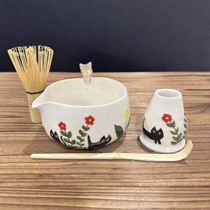 May include: A white ceramic matcha tea set with a whisk, bowl, and holder. The bowl and holder are decorated with black cat illustrations and red flowers. A small cat figurine sits on the bowl's rim. A bamboo scoop rests on a wooden surface.