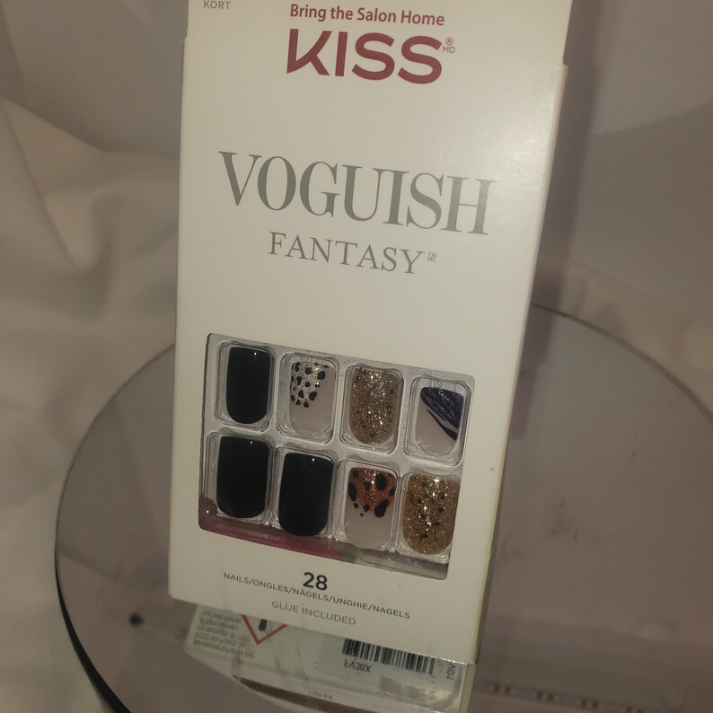 K&ouml;nnte beinhalten: Eine wei&szlig;e Schachtel mit KISS Voguish Fantasy Kunstn&auml;geln. Auf der Schachtel steht der Text "Bring the Salon Home" und "KISS" in Rot. Die N&auml;gel sind schwarz, goldglitzernd und mit Tiermustern und abstrakten Designs versehen. Die Schachtel enth&auml;lt 28 N&auml;gel.