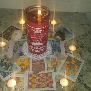 May include: A red candle with the words "Talisman Candle" and "Love Drawing Attract Amor" printed on it. The candle is surrounded by tarot cards and lit candles on a white countertop.