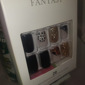 K&ouml;nnte beinhalten: Eine wei&szlig;e Schachtel mit Kunstn&auml;geln mit dem Wort "FANTASY" oben. Die N&auml;gel haben verschiedene Designs, darunter Schwarz, goldener Glitzer und Tiermuster. Die Packung enth&auml;lt 28 N&auml;gel.