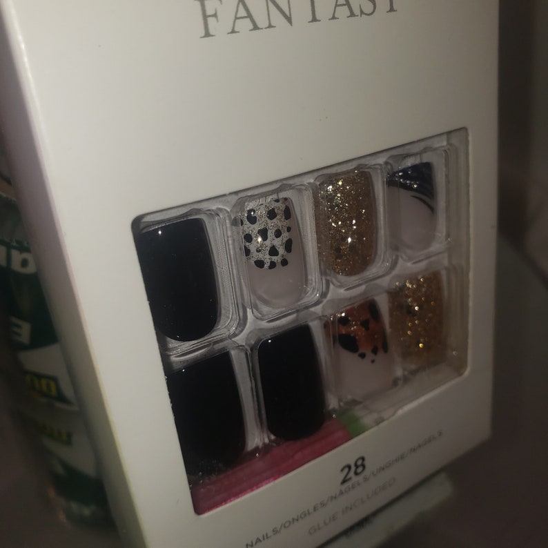 K&ouml;nnte beinhalten: Eine wei&szlig;e Schachtel mit Kunstn&auml;geln mit dem Wort "FANTASY" oben. Die N&auml;gel haben verschiedene Designs, darunter Schwarz, goldener Glitzer und Tiermuster. Die Packung enth&auml;lt 28 N&auml;gel.