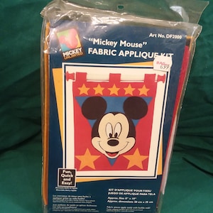 May include: A Mickey Mouse fabric applique kit featuring a red, white and blue castle banner. The kit includes fabric pieces, fusible web and instructions. The kit is approximately 20 cm by 25 cm.