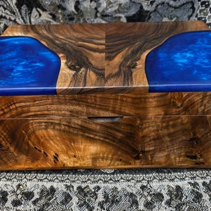 May include: A rectangular wooden box featuring a natural brown wood grain finish on the front and sides, with a lid that incorporates two large, vibrant blue resin sections on the left and right sides, framing a central piece of polished wood.