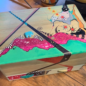 May include: Wooden box with a hand-painted abstract design. The box features a turquoise elephant figurine and a drawer. The artwork includes pink, green, and black elements, with a light wood finish. The box has metal corner protectors.