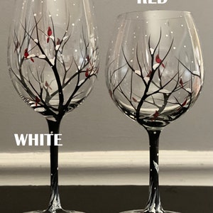 May include: Two hand-painted wine glasses with a winter scene. The glasses feature a black tree with white snow and red cardinals. One glass has a red stem and the other has a white stem. The words "RED" and "WHITE" are written on the image.