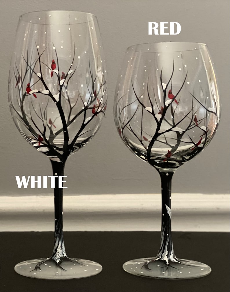 May include: Two hand-painted wine glasses with a winter scene. The glasses feature a black tree with white snow and red cardinals. One glass has a red stem and the other has a white stem. The words "RED" and "WHITE" are written on the image.
