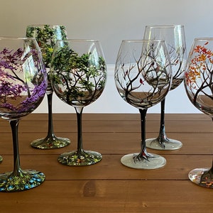 May include: Set of six hand-painted wine glasses with black stems and different colored trees on each glass. The trees are painted in shades of purple, green, brown, and orange.