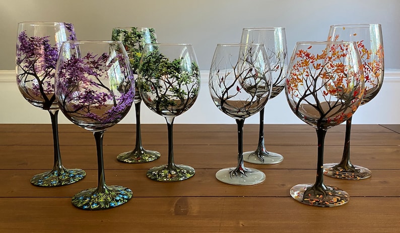 May include: Set of six hand-painted wine glasses with black stems and different colored trees on each glass. The trees are painted in shades of purple, green, brown, and orange.