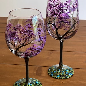 May include: Two hand-painted wine glasses with black stems and bases. The glasses feature a purple tree with blue flowers on the base.
