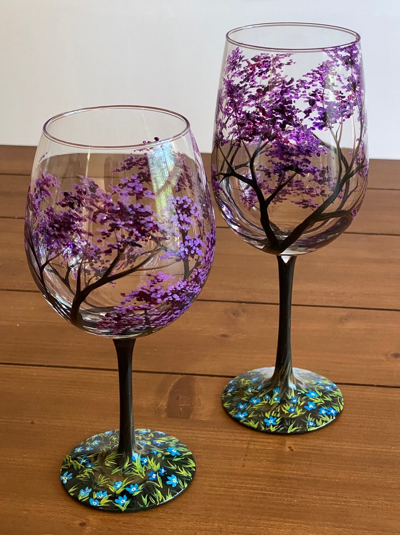 May include: Two hand-painted wine glasses with black stems and bases. The glasses feature a purple tree with blue flowers on the base.
