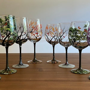May include: Set of six hand-painted wine glasses featuring trees with different colored leaves. The glasses have black stems and bases.
