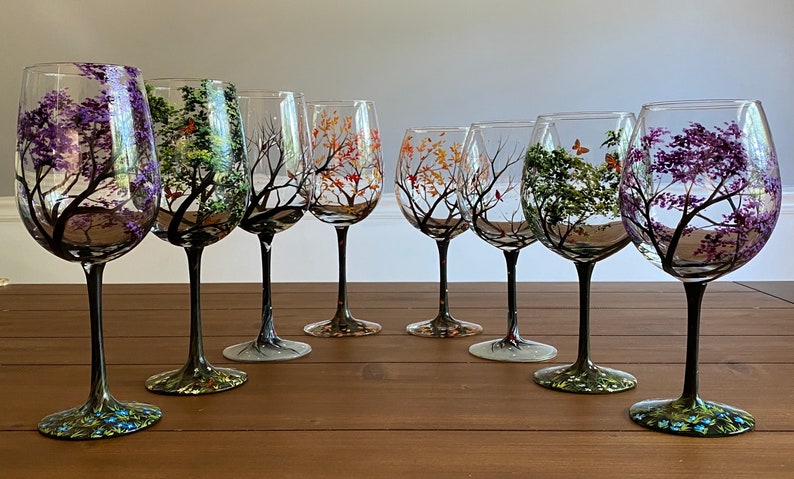May include: Set of six hand-painted wine glasses featuring trees with different colored leaves. The glasses have black stems and bases.