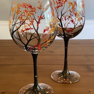 May include: Two clear glass wine glasses with black stems and bases. The glasses are hand-painted with a design of a tree with orange, yellow, and red leaves.