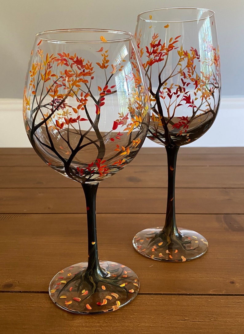 May include: Two clear glass wine glasses with black stems and bases. The glasses are hand-painted with a design of a tree with orange, yellow, and red leaves.