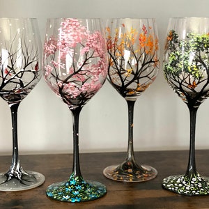 May include: Four wine glasses with hand-painted designs depicting the four seasons. The glasses feature a winter scene with snow, a spring scene with pink blossoms, a summer scene with green leaves, and a fall scene with orange leaves.