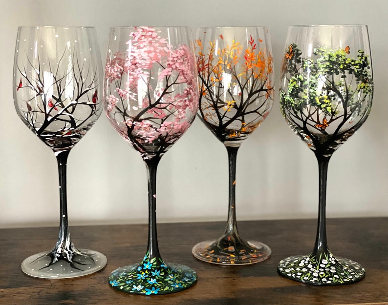 May include: Four wine glasses with hand-painted designs depicting the four seasons. The glasses feature a winter scene with snow, a spring scene with pink blossoms, a summer scene with green leaves, and a fall scene with orange leaves.