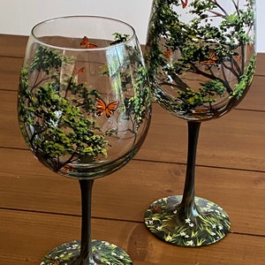 May include: Two hand-painted wine glasses with a forest scene and butterflies. The glasses have black stems and bases with a green and white floral design.