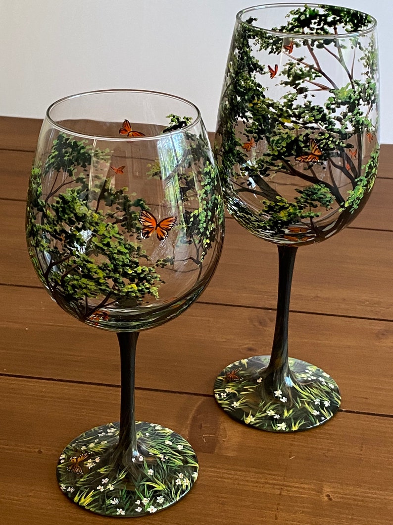May include: Two hand-painted wine glasses with a forest scene and butterflies. The glasses have black stems and bases with a green and white floral design.