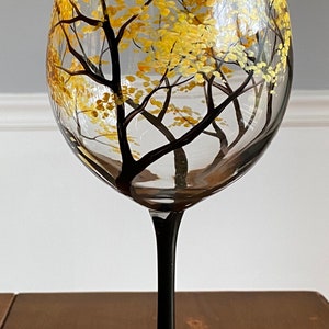 May include: A hand-painted wine glass with a black stem and base. The glass features a design of a forest scene with trees and yellow leaves. The base of the glass is painted with green grass and white flowers.