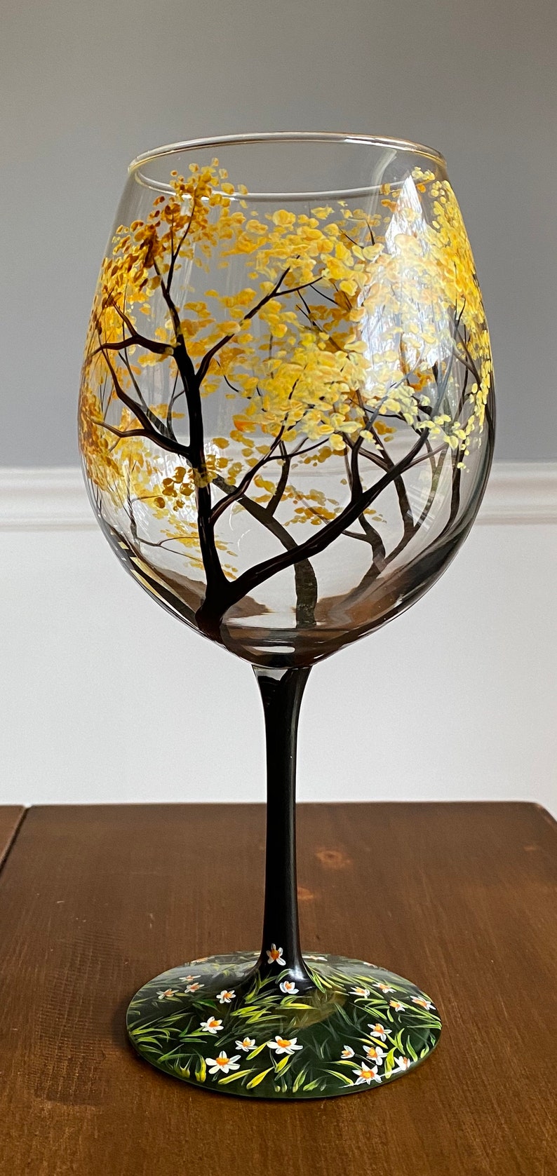May include: A hand-painted wine glass with a black stem and base. The glass features a design of a forest scene with trees and yellow leaves. The base of the glass is painted with green grass and white flowers.