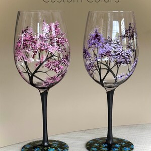 May include: Two hand-painted wine glasses with black stems and bases. The glasses feature a tree with pink and purple blossoms on the left and a tree with purple blossoms on the right. The bases of the glasses are decorated with blue flowers.