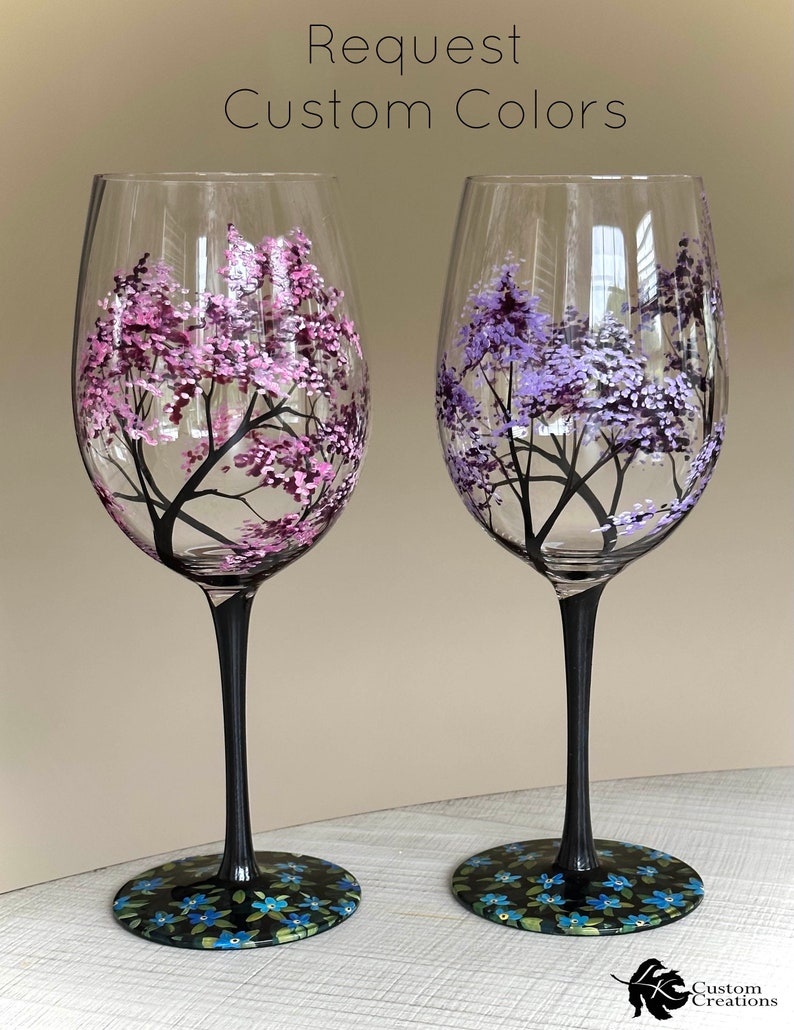 May include: Two hand-painted wine glasses with black stems and bases. The glasses feature a tree with pink and purple blossoms on the left and a tree with purple blossoms on the right. The bases of the glasses are decorated with blue flowers.
