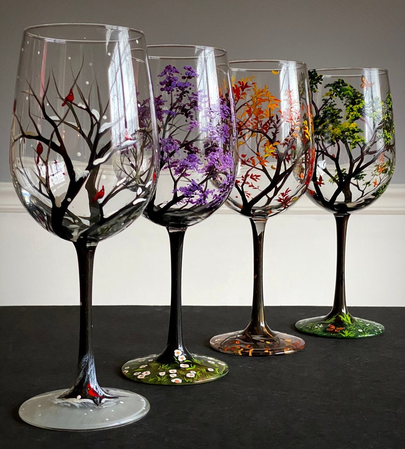 May include: Set of four hand-painted wine glasses, each depicting a different season. The glasses feature a black tree with colorful leaves and details representing each season: winter with snow and cardinals, spring with purple blossoms, summer with green leaves and a butterfly, and autumn with orange and yellow leaves.