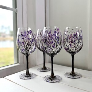 May include: Four clear wine glasses with a hand-painted design of a tree with purple leaves. The tree branches are black, and the stems and bases of the glasses are dark gray. The glasses are displayed on a white surface near a window.