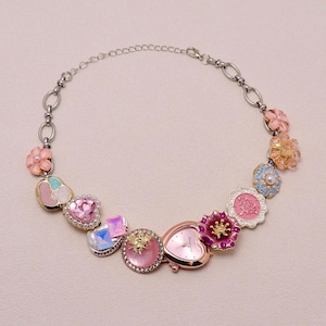 May include: A silver necklace with a heart-shaped watch and decorative charms. The charms include flowers, hearts, and gemstones in pink, blue, and iridescent colours. The watch face is pink.