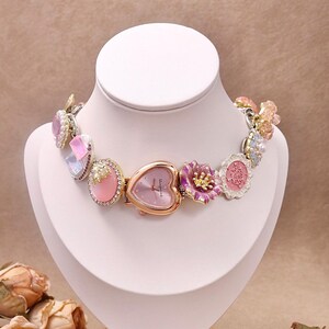 Op de afbeelding: Een decoratieve horlogeketting met een hartvormige wijzerplaat en bloemaccenten. Het horloge heeft een roze wijzerplaat en goudkleurig metaal. De ketting is versierd met roze, paarse en iriserende versieringen, tentoongesteld op een witte buste.