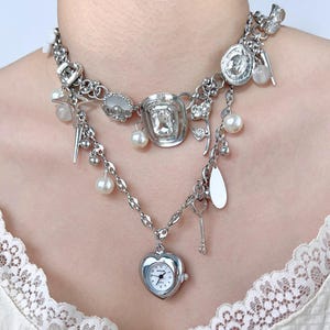 May include: A silver necklace featuring a heart-shaped watch pendant, pearls, and other charms. The layered design includes a key and other decorative elements. The necklace is a statement piece.