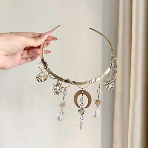 Op de afbeelding: Een goudkleurige choker-ketting met hemelse bedels. De ketting heeft zon-, maan- en kristaldruppelhangers. Een schelpenbedel is ook inbegrepen. De ketting wordt door een hand tegen een neutrale achtergrond gehouden.