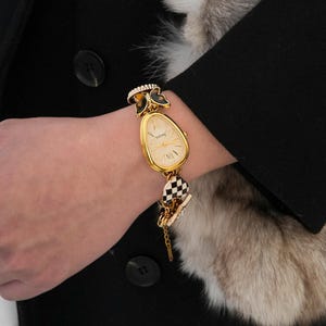Op de afbeelding: Een goudkleurig polshorloge met een crèmekleurige wijzerplaat en zwarte Romeinse cijfers. Het horloge heeft een unieke druppelvorm en is versierd met zwarte vlinderaccenten en een zwart-wit geblokte bedel. Het horloge zit om een pols.