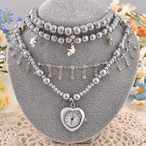 Op de afbeelding: Een set van drie strengen grijze parelkettingen op een grijze mannequin. De bovenste twee strengen hebben sterbedels. De onderste streng heeft een hartvormige horlogehanger. Strass accenten en bungelende bedels voegen details toe. Bloemenaccenten zijn op de achtergrond.
