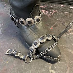 May include: Black leather cowboy boot with a silver chain and silver concho accents. The boot has a pointed toe and a high heel.