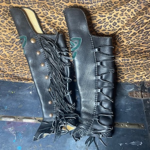 May include: A pair of black leather chaps with decorative fringe, brass-colored studs, and green Celtic knot accents. The chaps are designed with a lace-up closure on one side and are displayed on a leopard print background.