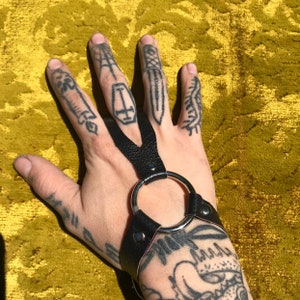 May include: Black leather fingerless gloves with a silver ring and a black leather strap. The gloves have a V-shaped opening between the index and middle fingers. The gloves are worn on a hand with several tattoos.