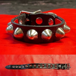 May include: A black leather bracelet with silver pyramid-shaped studs. The bracelet has a buckle closure.