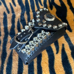 May include: A black leather wallet with silver studs and a buckle closure. The wallet has a unique design with a flap that folds over the front.