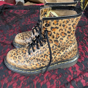 May include: A pair of leopard print combat boots with black laces and yellow pull tabs. The boots have a classic design with a round toe and a sturdy sole. The animal print pattern features brown and black spots.